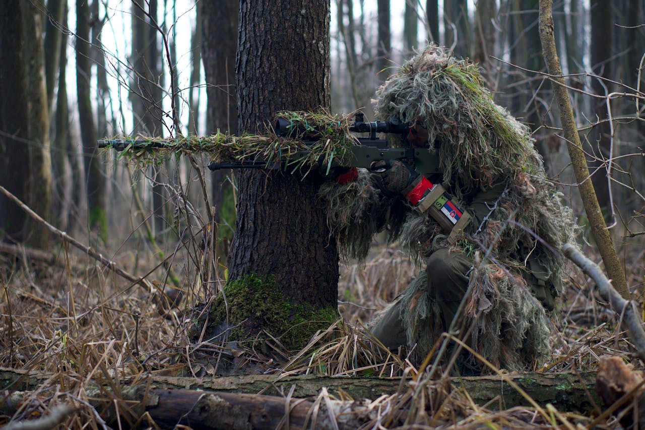 Sniper in camouflage gear taking aim in a dense forest environment.